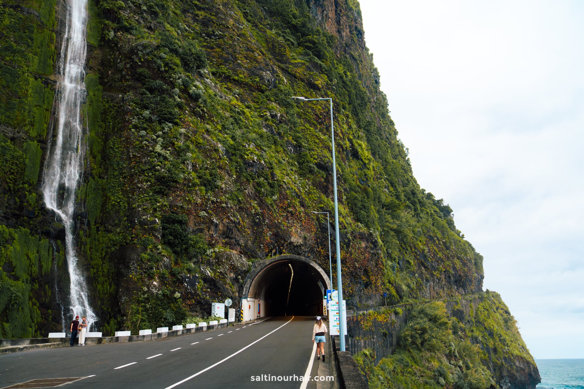 A road tunnel cuts through a steep, green cliffside with a waterfall flowing down beside it; a person stands near the tunnel entrance, exploring one of the best hikes Madeira has to offer.