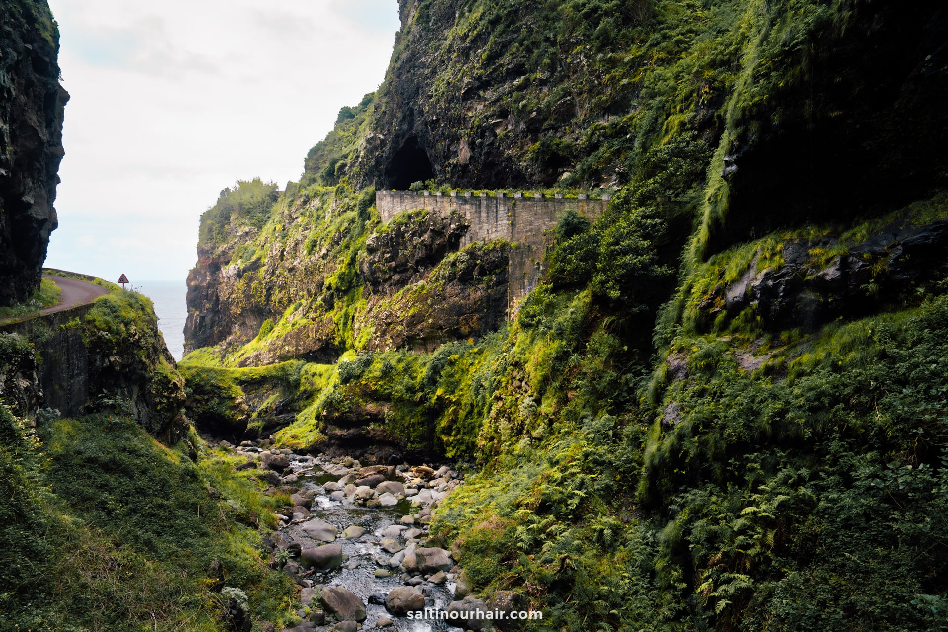 Narrow road and tunnel carved into steep, rocky green cliffs above a rocky stream, with a person walking in the distance — Ribeira do Inferno (ER101) — a glimpse of the best hikes Madeira has to offer.