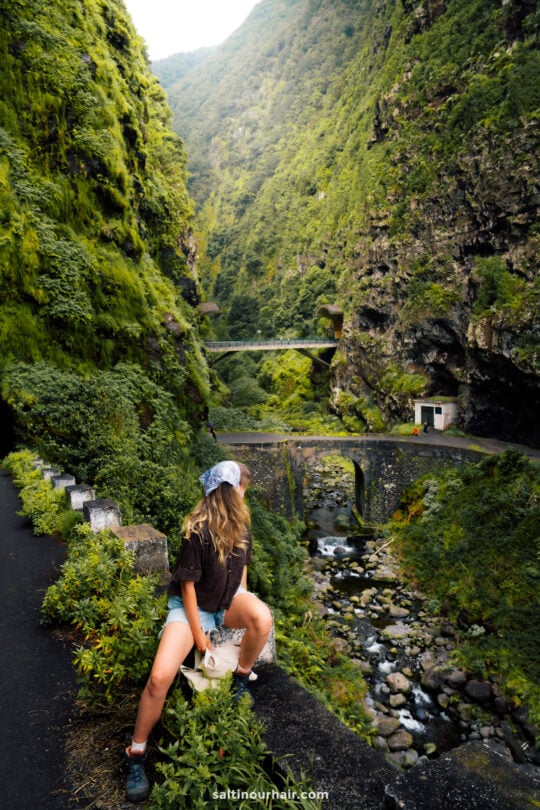 A person with long hair and a headscarf sits on a rock beside a winding mountain road called 