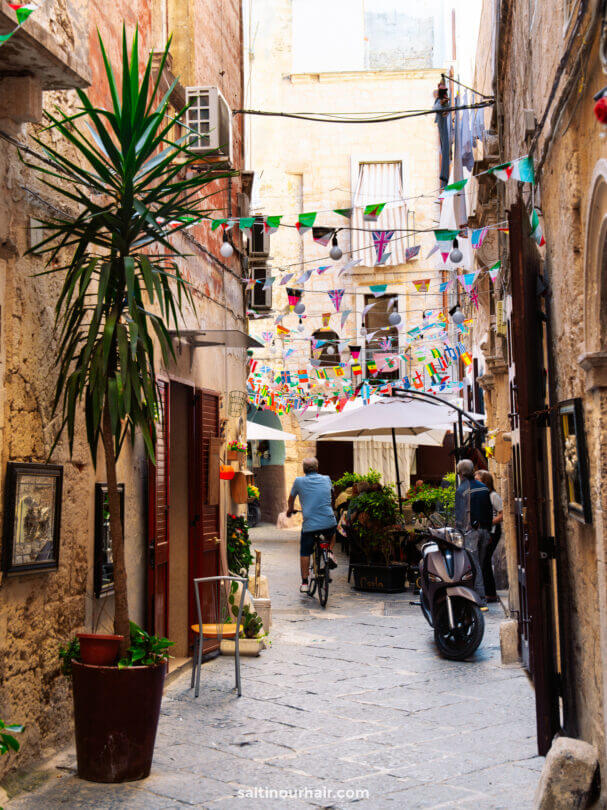 A narrow, cobblestone alley in Bari, Puglia, with potted plants, hanging flags, outdoor seating, a parked scooter, and a person riding a bicycle 