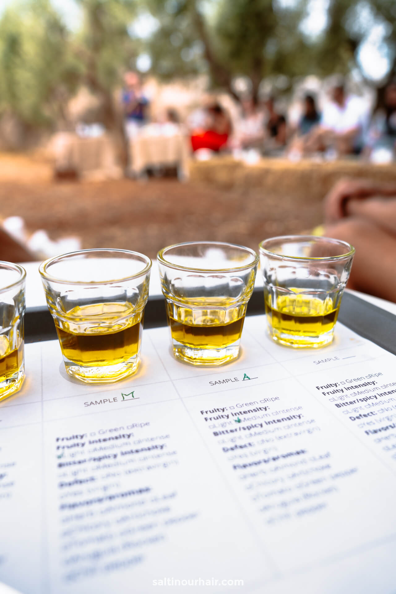 Four small glasses of olive oil are arranged on a tray over a tasting sheet, set outdoors in Puglia, Italy, with blurred people in the background.