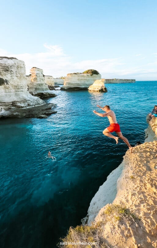 A man jumps off a rocky cliff into the clear blue water at Marina de Ostuni, puglia italy while another person swims below. - the perfect day trip from ostuni puglia italy