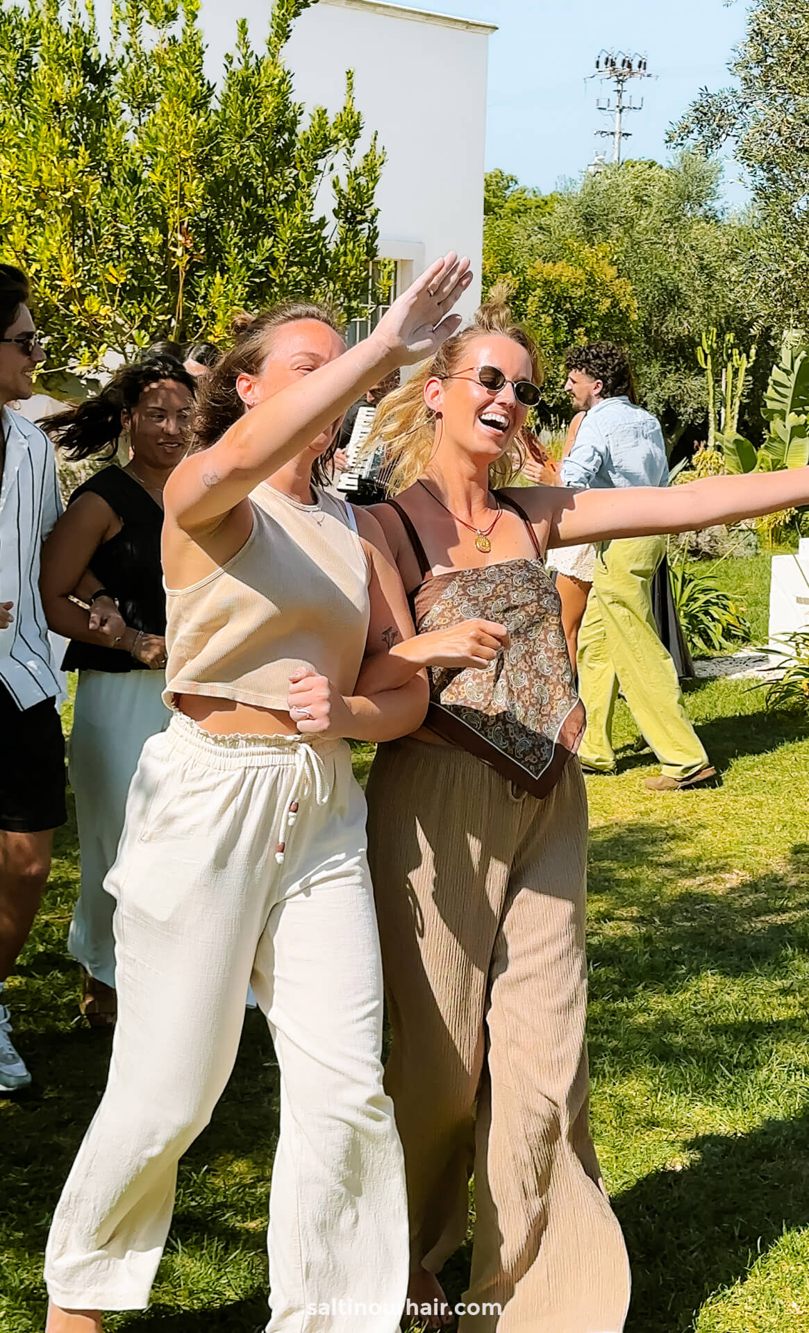 Two women dancing outdoors on grass in Puglia, Italy, smiling and waving, surrounded by other people 