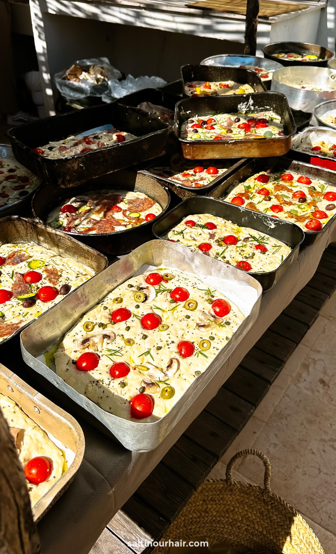 Multiple trays of unbaked focaccia are topped with cherry tomatoes and herbs and arranged on a wooden table in bright natural light for a cooking class in Puglia Italy