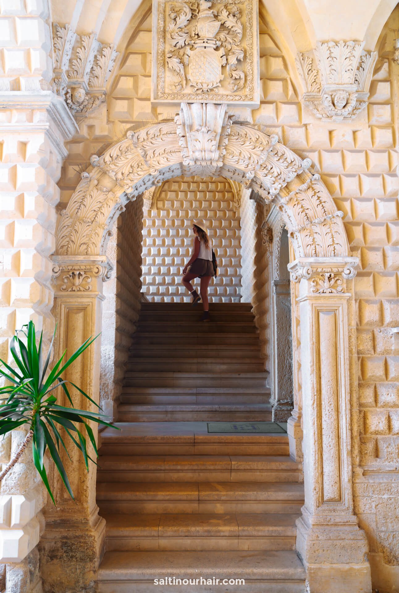 A woman walks up stone stairs through an ornate arched entryway with detailed stonework and carvings in Puglia, Italy