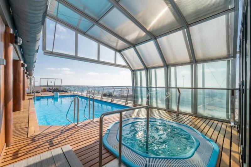 Indoor pool area at ARTIEM Capri in Menorca, featuring a hot tub, wooden deck, and expansive glass windows and ceiling with breathtaking views of the sky and horizon.