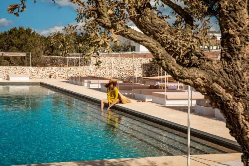 A person in a yellow outfit and wide-brimmed hat sits at the edge of a clear swimming pool at Menorca Experimental Hotel, with a tree in the foreground and stone walls in the background.