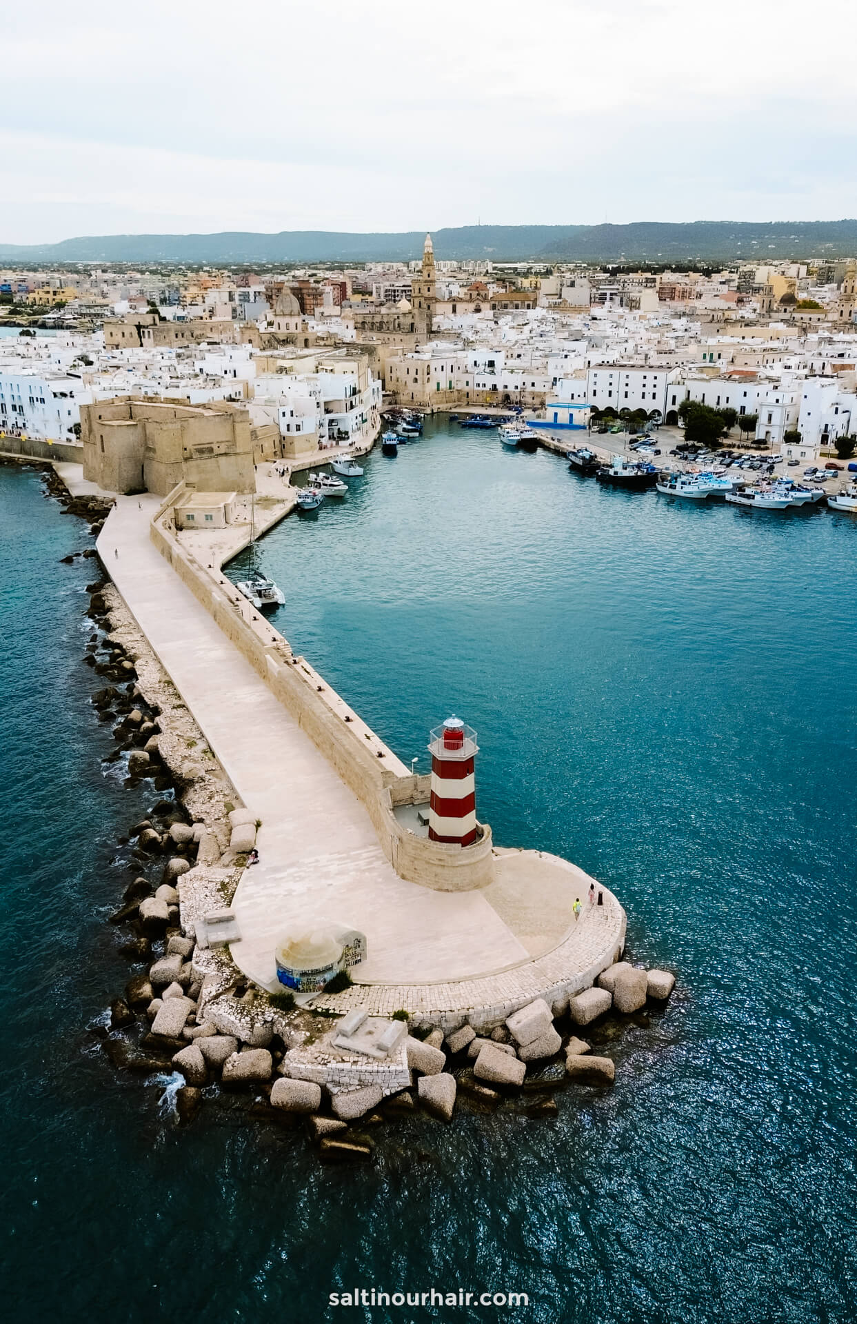 Aerial view of a red and white striped lighthouse on a stone pier extending from the Lungomare into a harbor of monopoli puglia italy, with a historic coastal town and boats in the background.