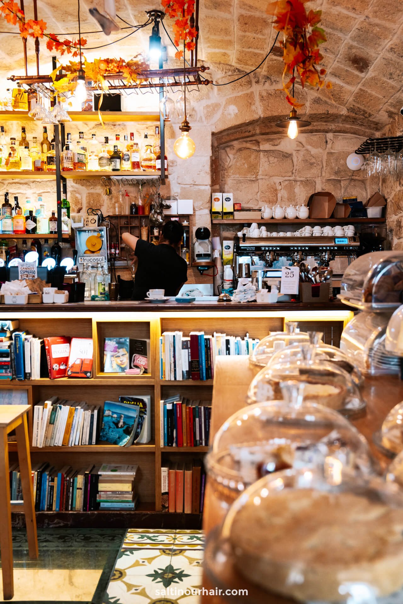 A cozy café in Monopoli, Italy, with a stone ceiling, bookshelves, and a counter displaying covered pastries; a person works behind the bar stocked with bottles and glasses.