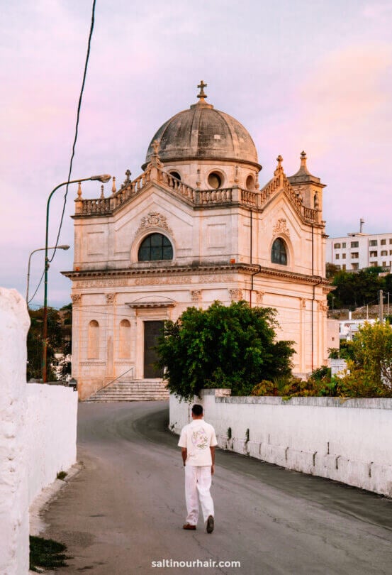 A person in white clothing walks down a curved road toward a large, domed stone building with arched windows under a pastel sky in Puglia, Italy.