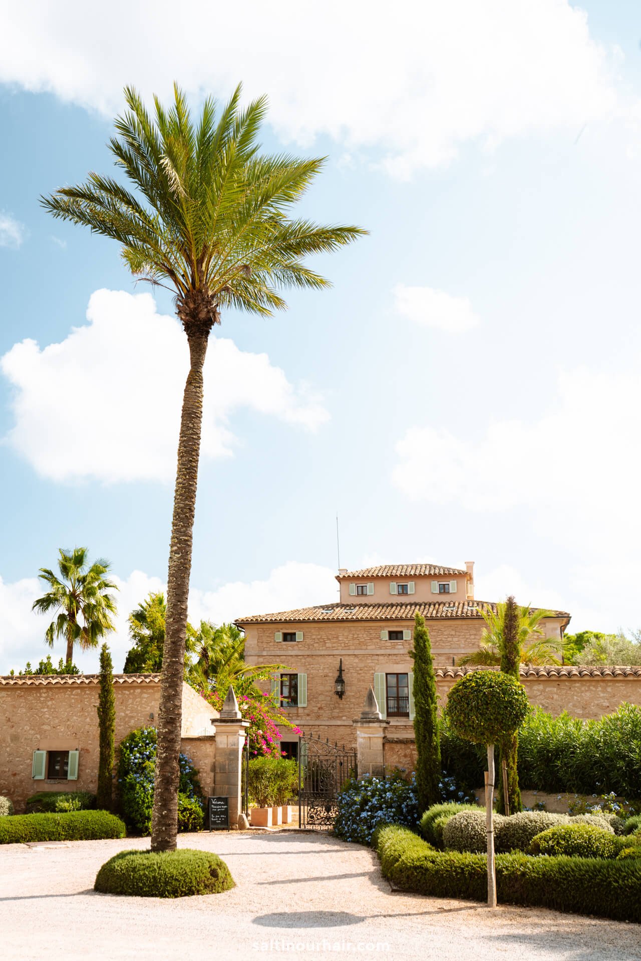 A Mediterranean-style villa with stone walls, surrounded by manicured gardens and palm trees, sits under a bright blue sky with scattered cloudsâ€”perfect for a restaurant Santanyi Mallorca experience.