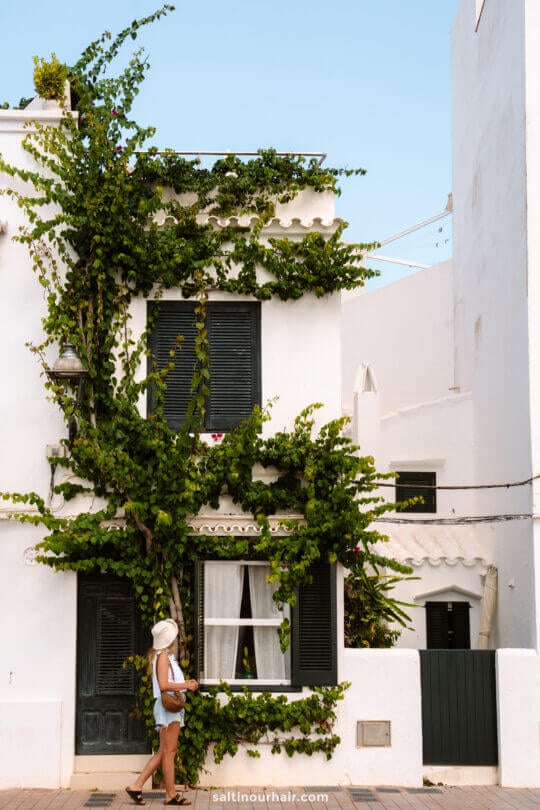 A woman in a sunhat walks past a white two-story house with black shutters and lush green vines, evoking the charm of Menorca in the Balearic Islands.