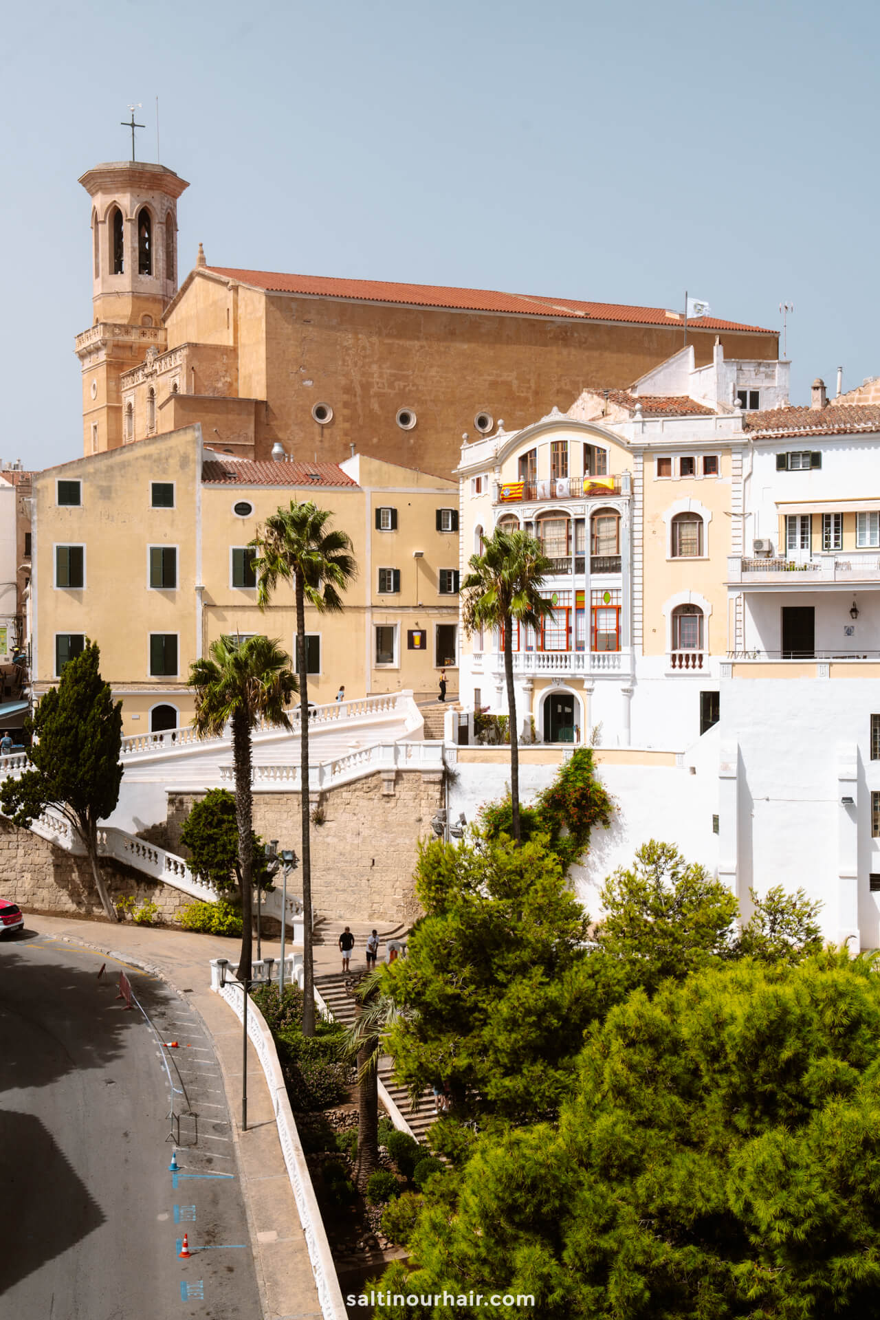 View of a Mediterranean town in Menorca, Balearic Islands, with historic buildings, palm trees, and a large church in the background under a clear sky.