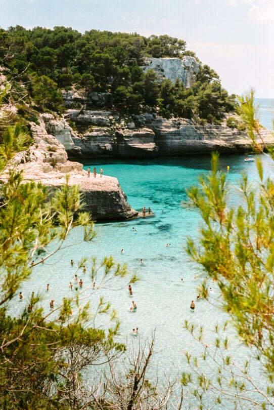 People swim and relax in a clear turquoise bay surrounded by rocky cliffs and pine trees on a sunny day in Menorca, one of the stunning Balearic Islands.