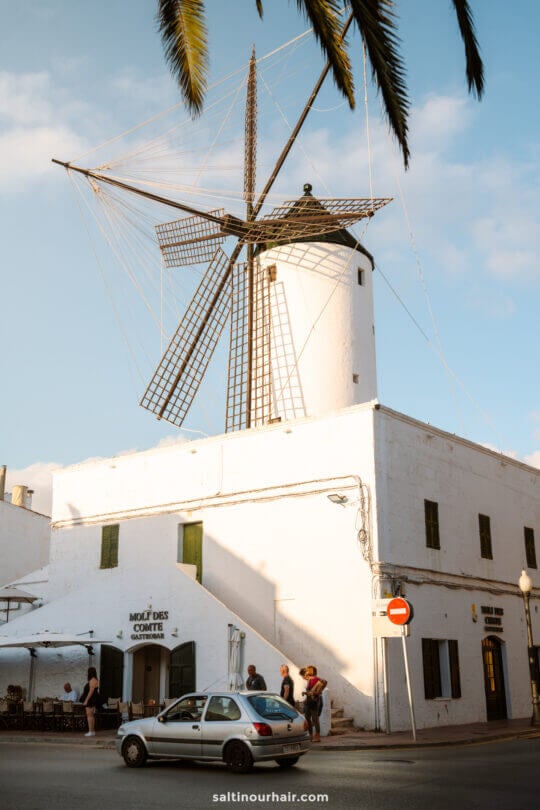 A traditional white windmill with large blades stands on top of a white building with green window shutters in Menorca, Balearic Islands; people and a car are in front of the building.