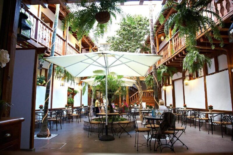 Open-air courtyard restaurant at Hotel Emblemático Tenerife, featuring wooden balconies, hanging ferns, large white umbrellas, and tables where people are dining.