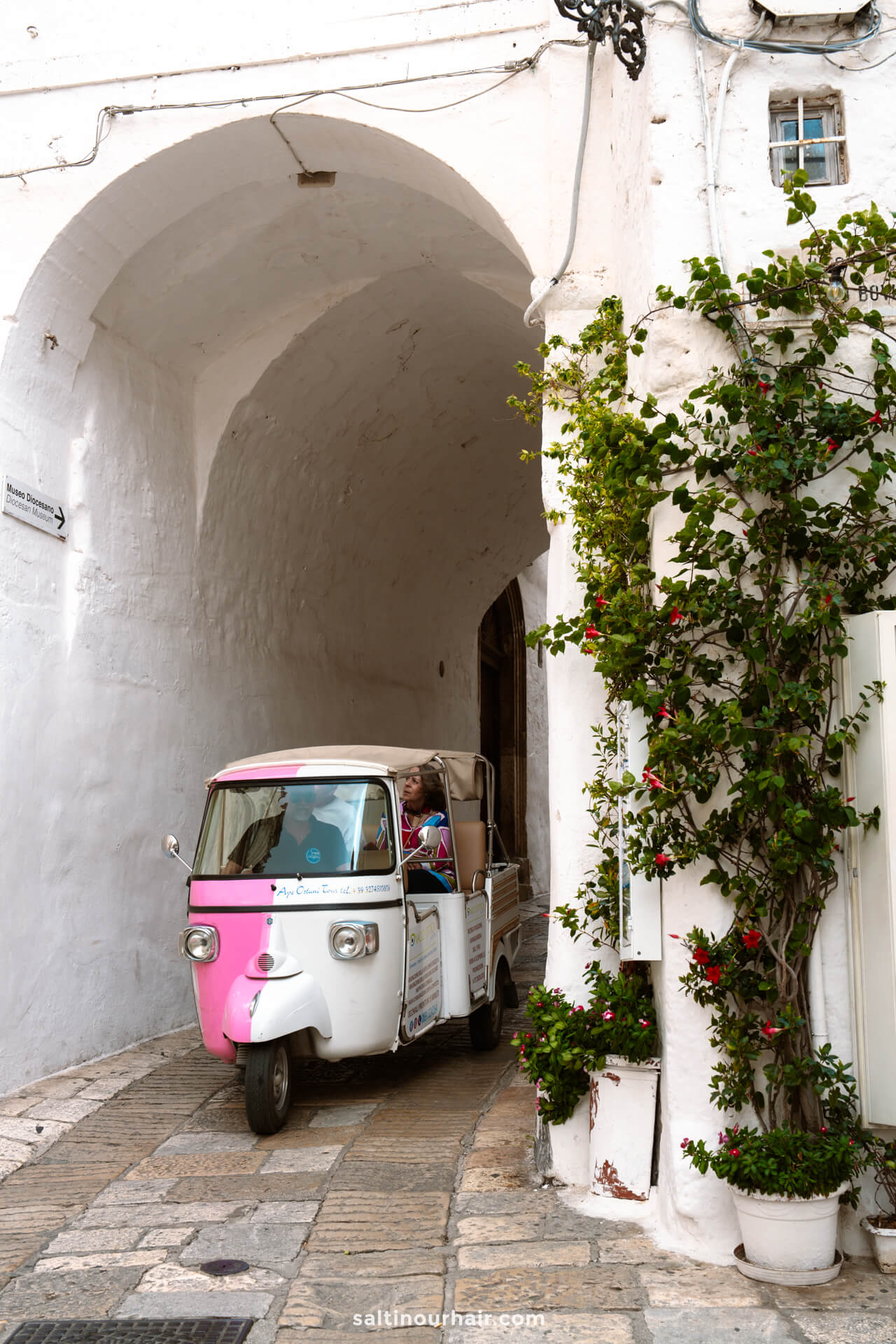 A white and pink tuk-tuk with two passengers drives through a narrow, arched stone street in Ostuni, puglia italy lined with white buildings and green vines.