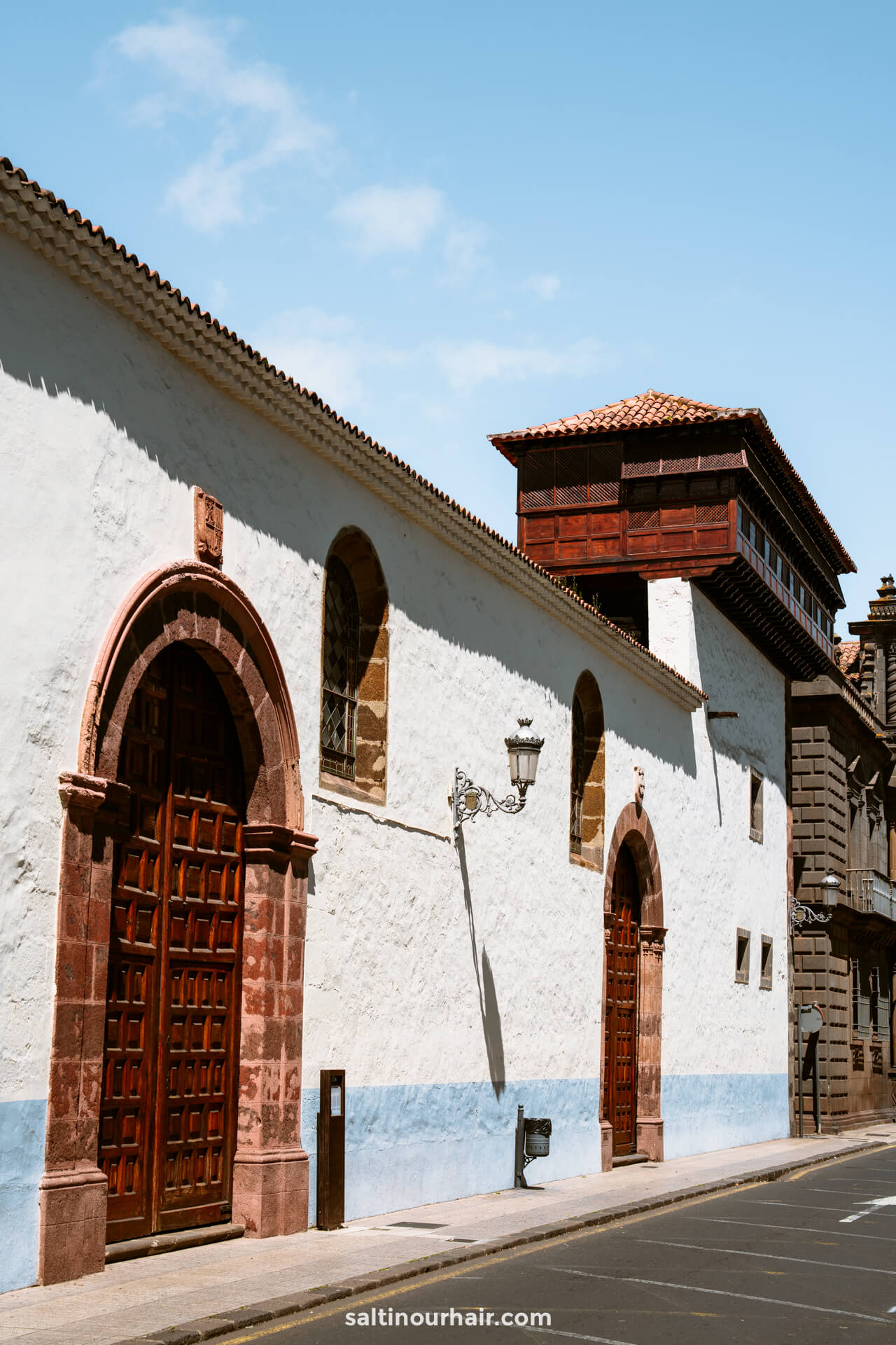 A white stucco building with red stone archways, wooden doors, and a balcony, nestled along a quiet street under a blue sky in San Crist&oacute;bal de La Laguna, Tenerife.