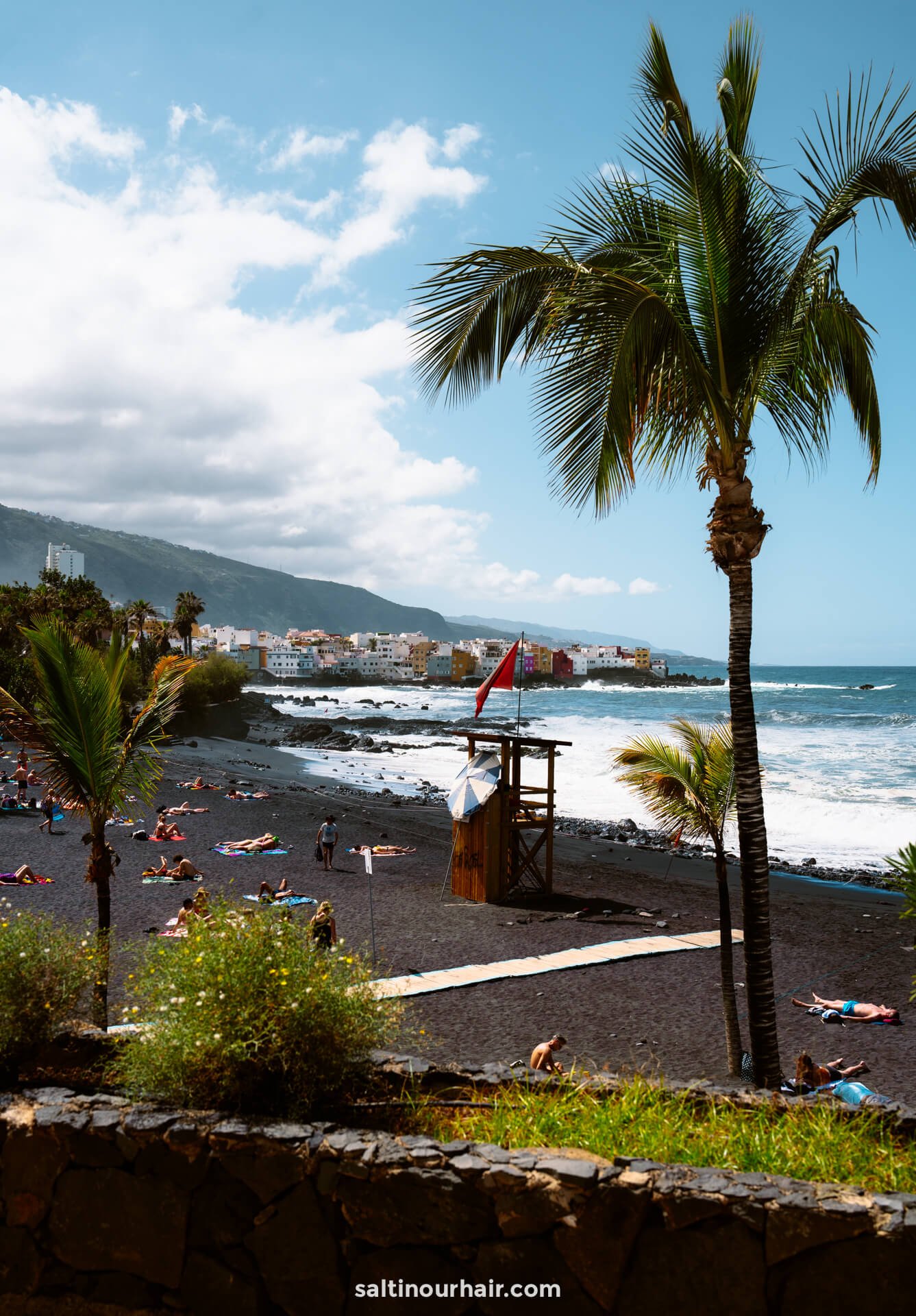 People sunbathe on Playa Jardin in Puerto de la Cruz, Tenerife, a black sand beach with palm trees, gentle waves, and colorful buildings in the distance under a partly cloudy sky.