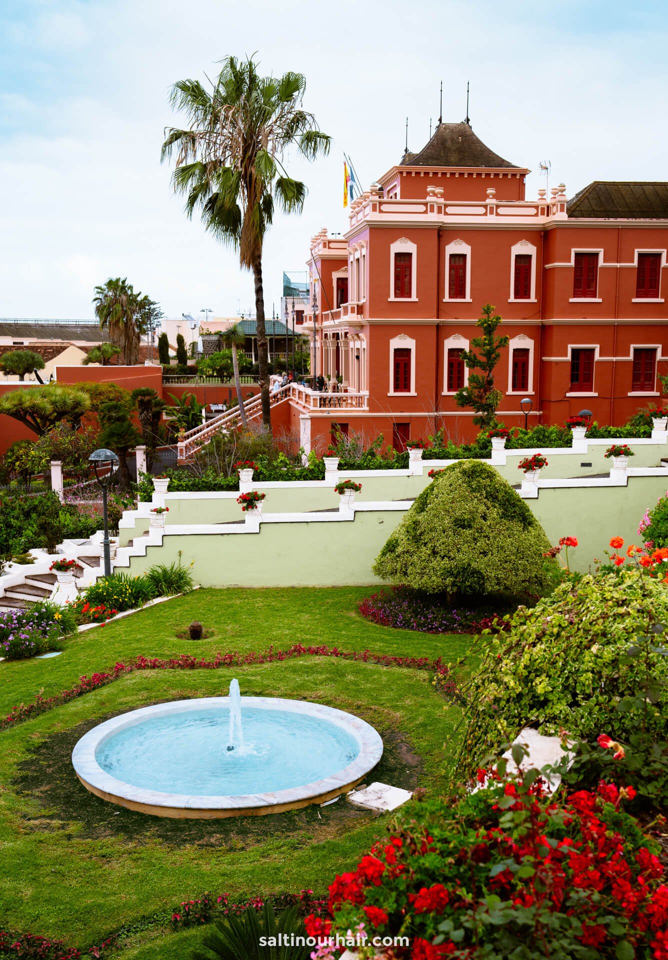 Marquesado de la Quinta Roja Garden with red Club Social Liceo Taoro building in the background under a partly cloudy skyâ€”one of the charming things to do in La Orotava.