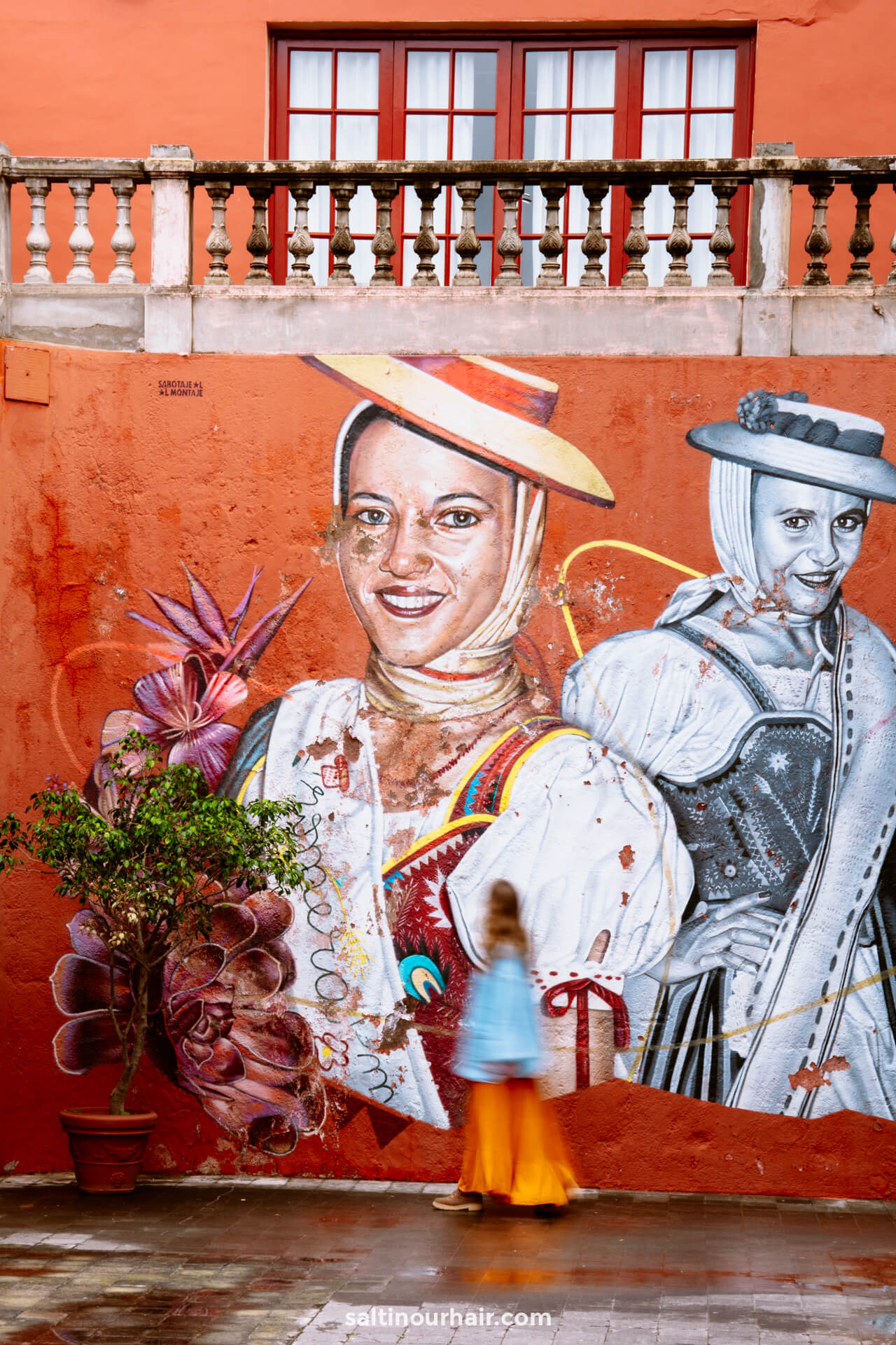 A mural depicting two women in traditional attire is painted on an orange wall; a blurred person in colorful clothing walks past a potted plantâ€”capturing the vibrant street culture among the many things to do Tenerife offers.