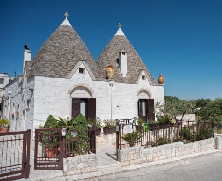 Charming white stone house with two conical roofs in Alberobello’s Grandi Trulli style. Features a gated front yard, lush plants, pottery on the roof, and a serene blue sky—an inviting Bed & Breakfast escape.
