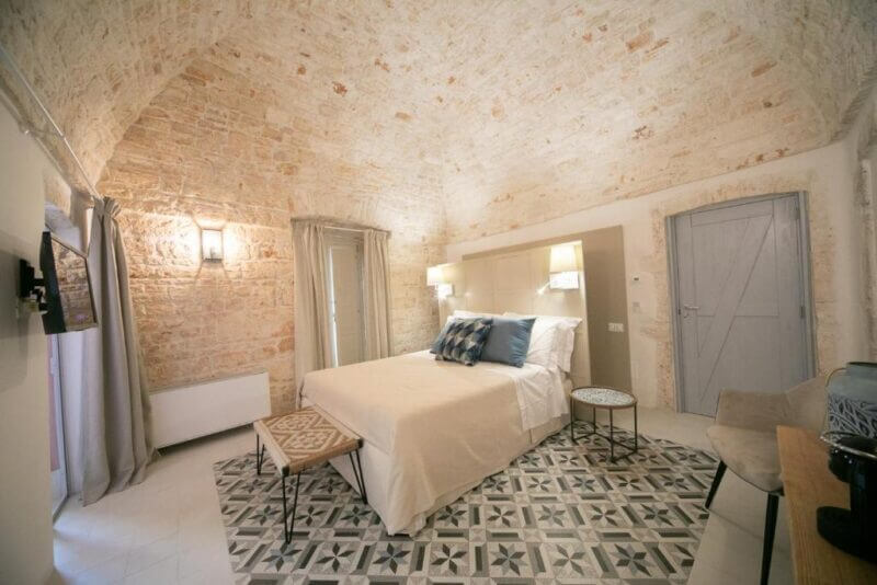 A Boutique Room in Alberobello featuring a vaulted stone ceiling, neutral décor, patterned tile floor, double bed with pillows, side tables, and a light blue door.
