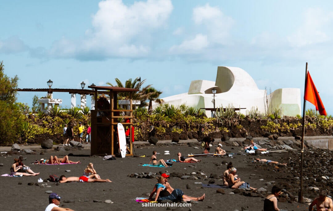 People sunbathing on the black sand of Playa Jardin, one of the top things to do in Puerto de la Cruz, Tenerife, with a lifeguard tower, red flag, and modern white building in the background.