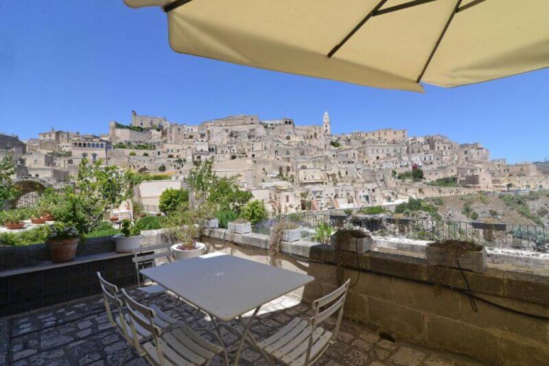 A patio at La Corte Dei Pastori with a white table and chairs under an umbrella overlooks the ancient stone hillside town of Matera beneath a clear blue sky.