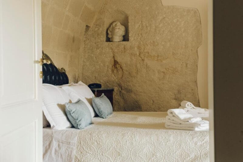 A neatly made bed with white and blue pillows faces a stone wall with a small statue in a niche; folded towels are placed at the foot of the bed, capturing the cozy elegance of Casastella Hotel in Matera.