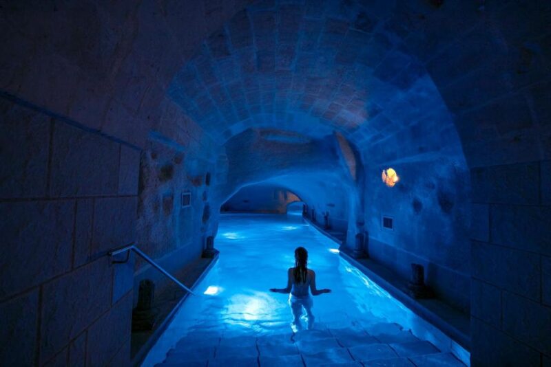 Person standing in waist-deep water inside a dimly lit, blue-illuminated stone tunnel with arched ceiling at Matera’s Locanda Di San Martino hotel.