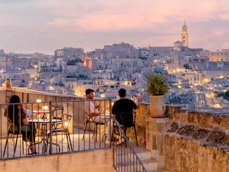 People sit at outdoor tables on the Il Belvedere terrace overlooking Matera’s historic hillside town at sunset, as city lights begin to illuminate the ancient stone buildings.
