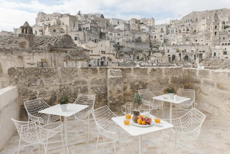 Outdoor terrace at Dimora Di Metello features white wire chairs and tables set with fruit, juice, and plants, overlooking Matera’s historic stone buildings on a hillside under a partly cloudy sky—a perfect Matera accommodation experience.