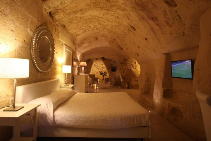 A modern Hotel bedroom in Matera with stone cave walls and ceiling, a neatly made bed, white furniture, and a wall-mounted TV. The room is softly lit by bedside lamps, capturing the unique Caveoso charm.