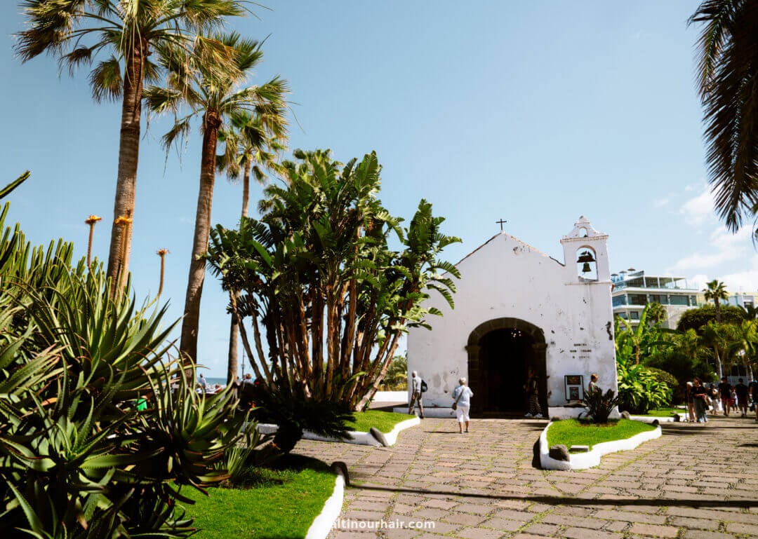 Whitewashed church in Puerto de la Cruz, Tenerife, surrounded by palm trees and lush greenery, with people walking along a stone pathway beneath the blue sky.