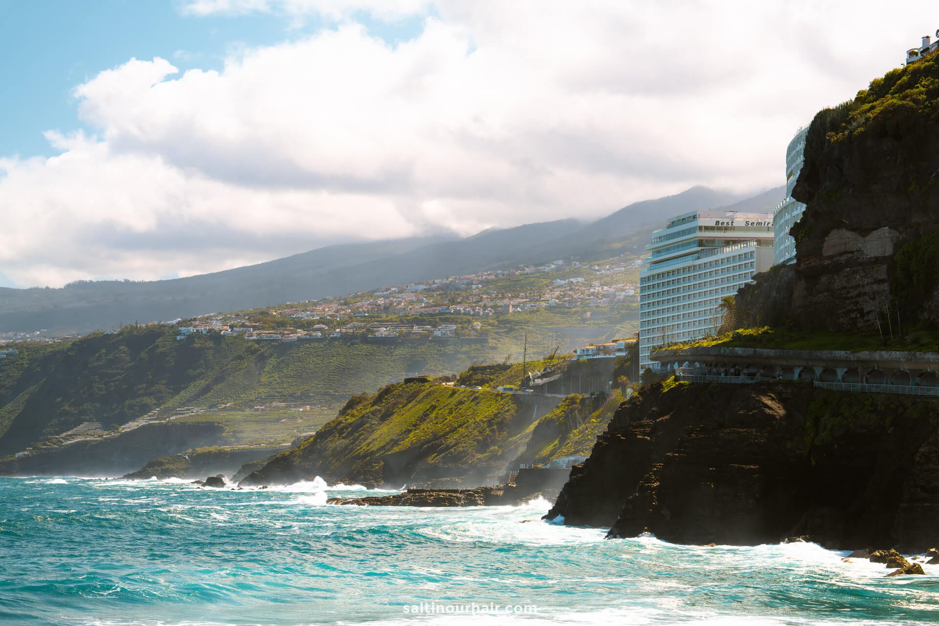A seaside cliff with waves crashing below, modern buildings on the cliffside, and rolling hills in the background under a partly cloudy skyâ€”much like the scenic views near Playa JardÃ­n, one of the top things to do in Puerto de la Cruz, Tenerife.