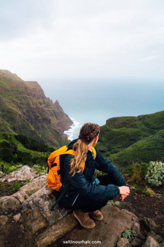 woman with a yellow backpack and braided hair sits on a rock, overlooking the green mountainous landscape and ocean of Anaga Rural Park, the perfect day trip from San Crist&oacute;bal de La Laguna
