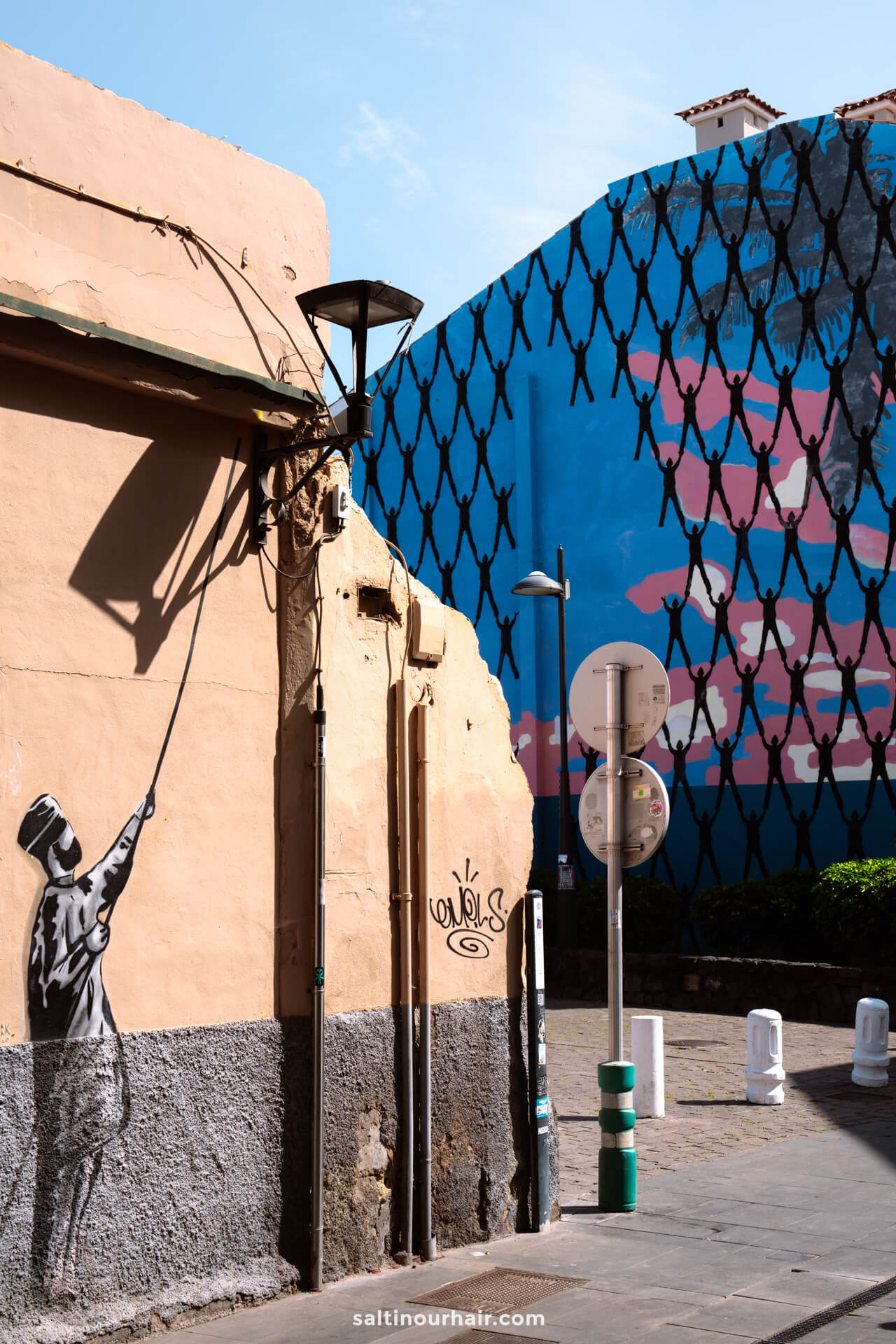 Street art of a person painting up a wall is seen on a beige building in La Ranilla Neighborhood, Puerto de la Cruz, Tenerife; in the background, a blue mural with netted patterns covers another building.