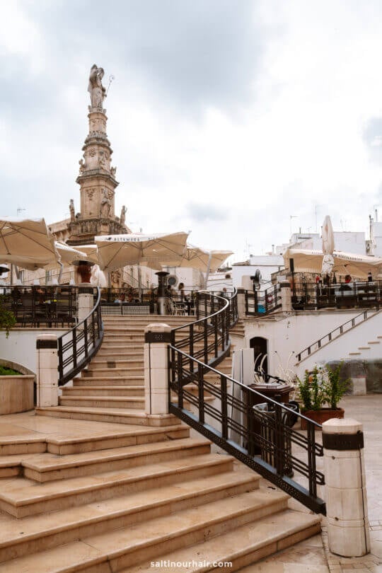 Stone steps lead up to an outdoor cafe with tables and umbrellas in Ostuni; a tall ornate statue stands in the background under a cloudy sky. A perfect stop on your puglia itinerary
