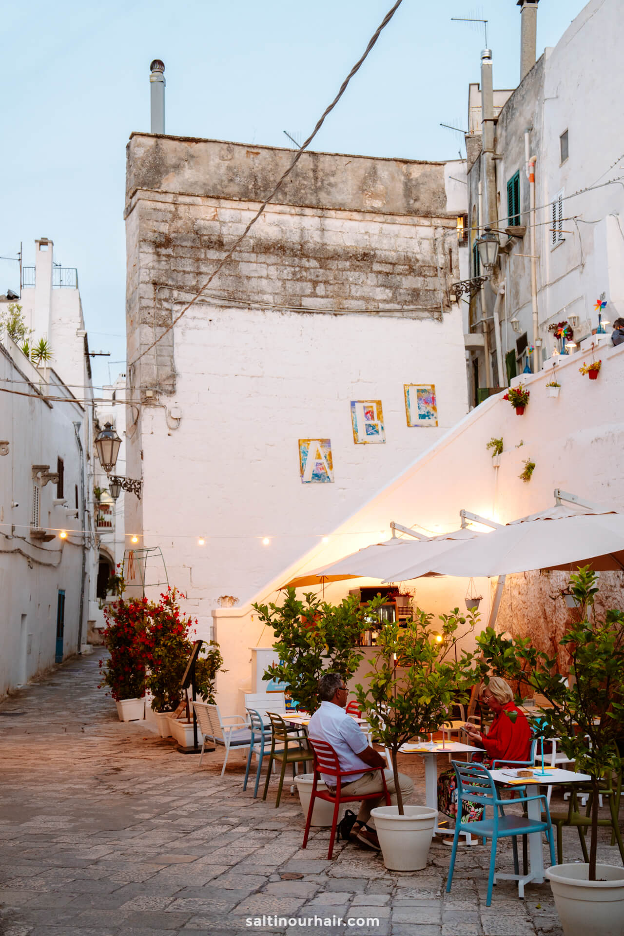 A small outdoor café with tables and chairs sits beside Ostunis whitewashed buildings on a quiet cobblestone street.