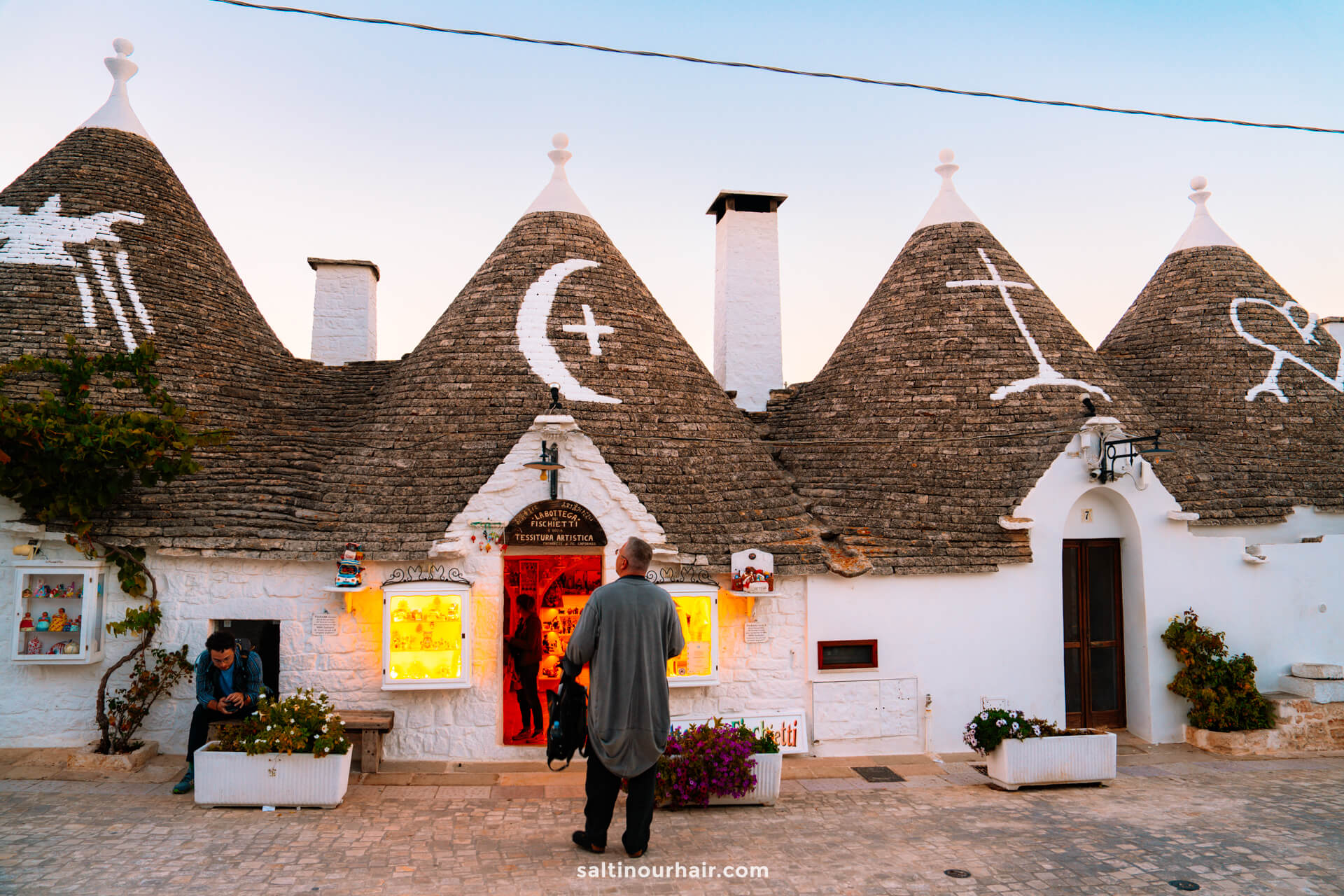 A person stands facing a white stone trulli houses in Alberobello, Italy, each marked with a large white symbol. Warm light glows from a shop window, while another person sits to the left on the steps.