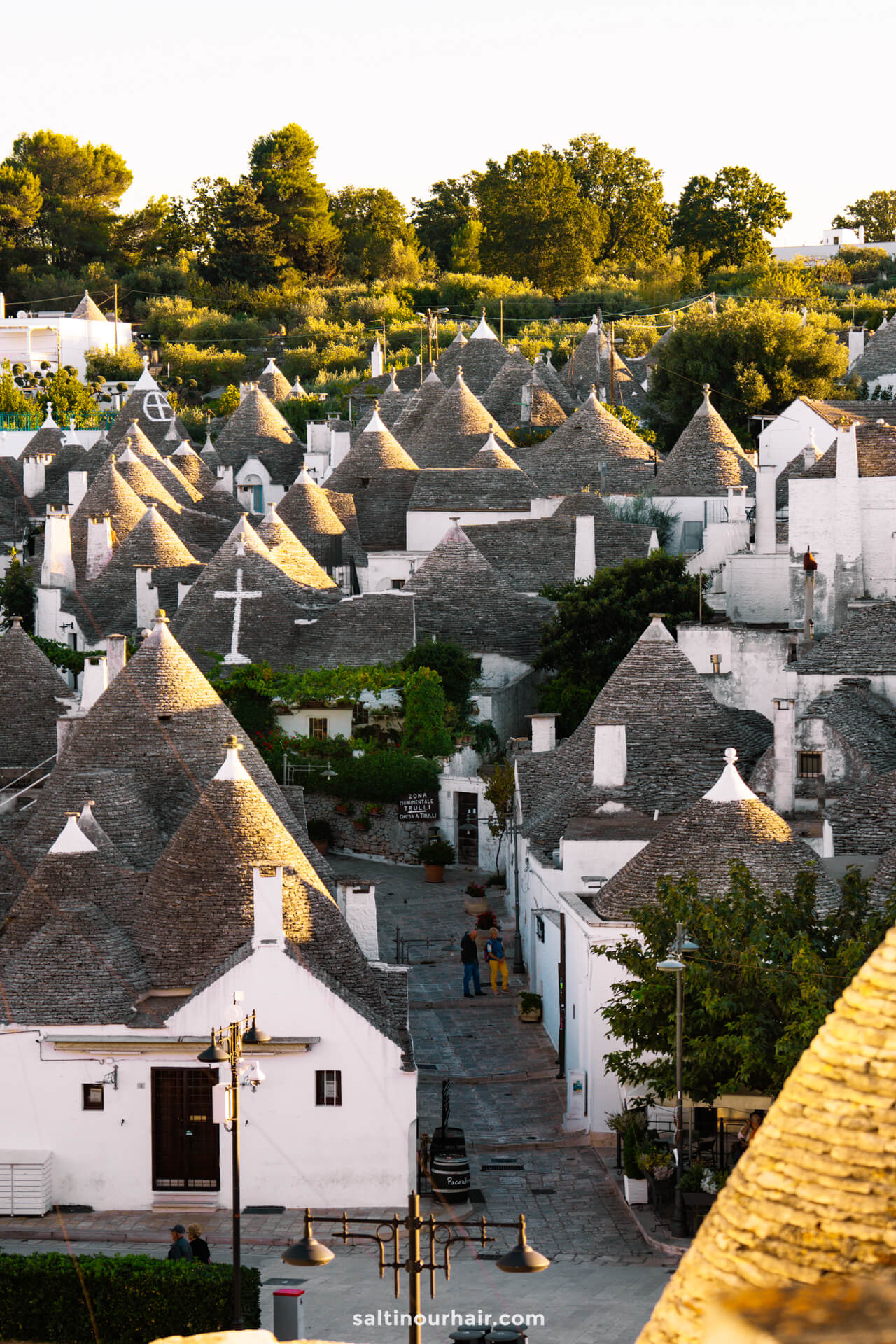 A scenic view of Alberobello, Italy, featuring its traditional whitewashed houses with conical stone roofs, framed by lush greenery at sunset. Discover the timeless charm that makes Alberobello Italy truly unique.
