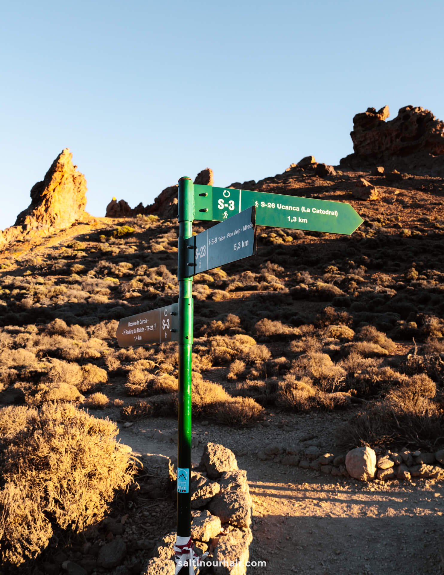 A green hiking trail signpost with multiple directional arrows stands on a rocky, shrub-covered landscape in Teide National Park under a clear sky at sunset.