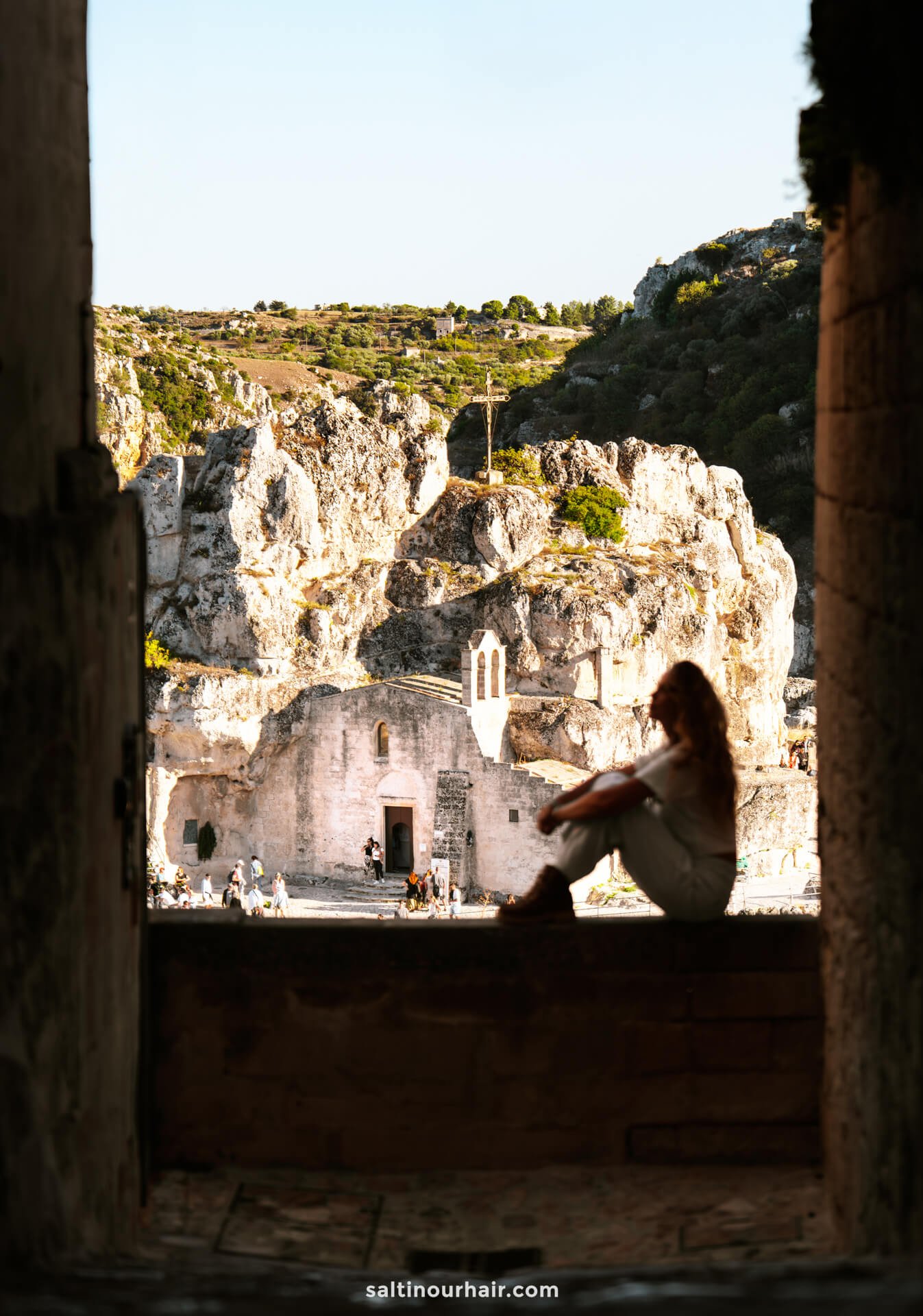 A woman sits in shadowed foreground, looking out towards an ancient stone building and the rocky landscape of Matera, Italy