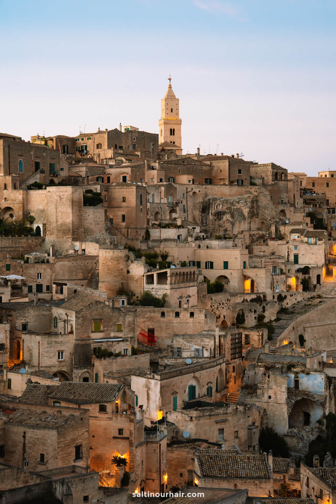 A hillside view of stone buildings with small windows and a tall church tower at the top, captured at dusk in the historic cityscape of Matera, Italy.