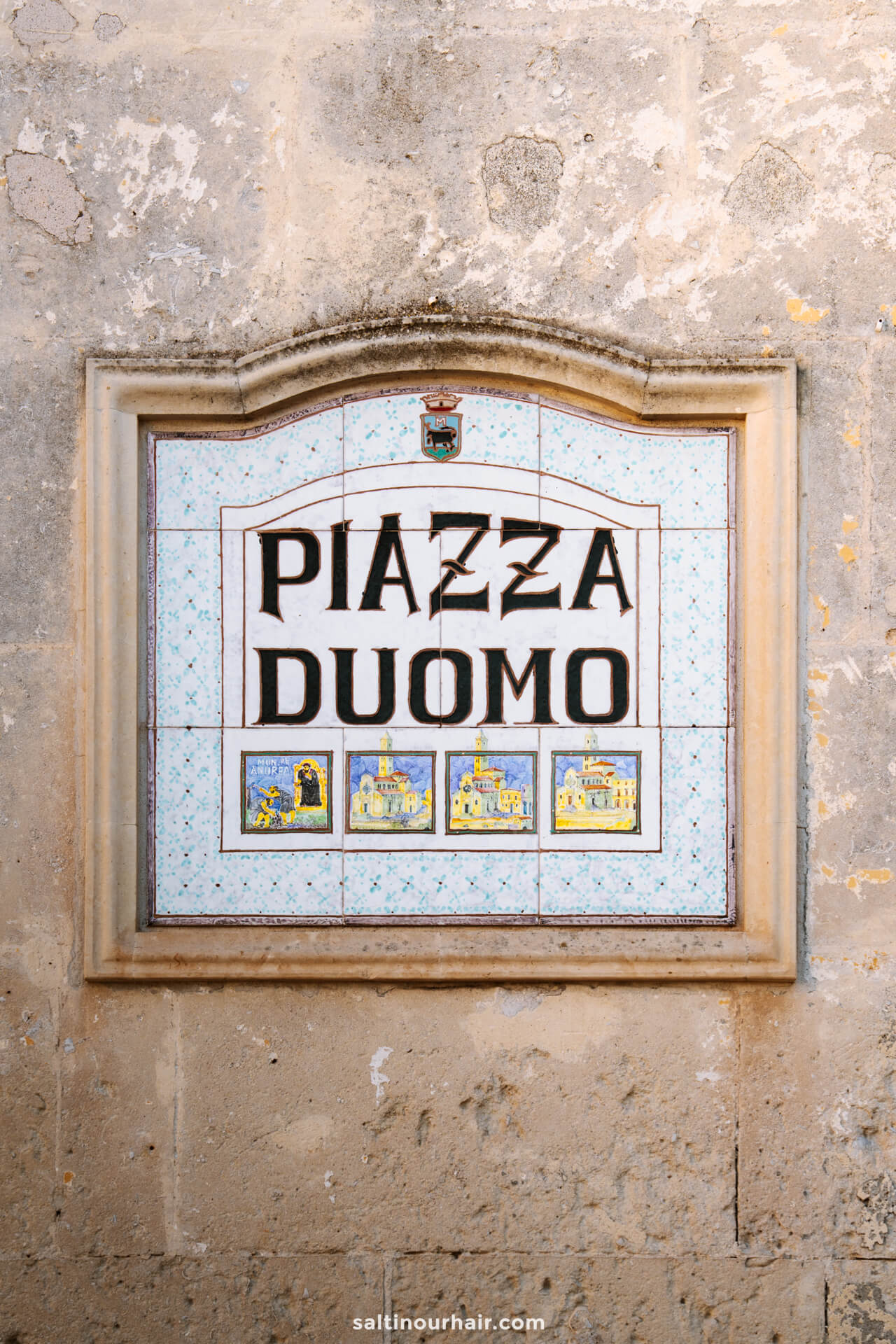 A ceramic street sign reading Piazza Duomo, matera with small illustrated tiles depicting buildings.