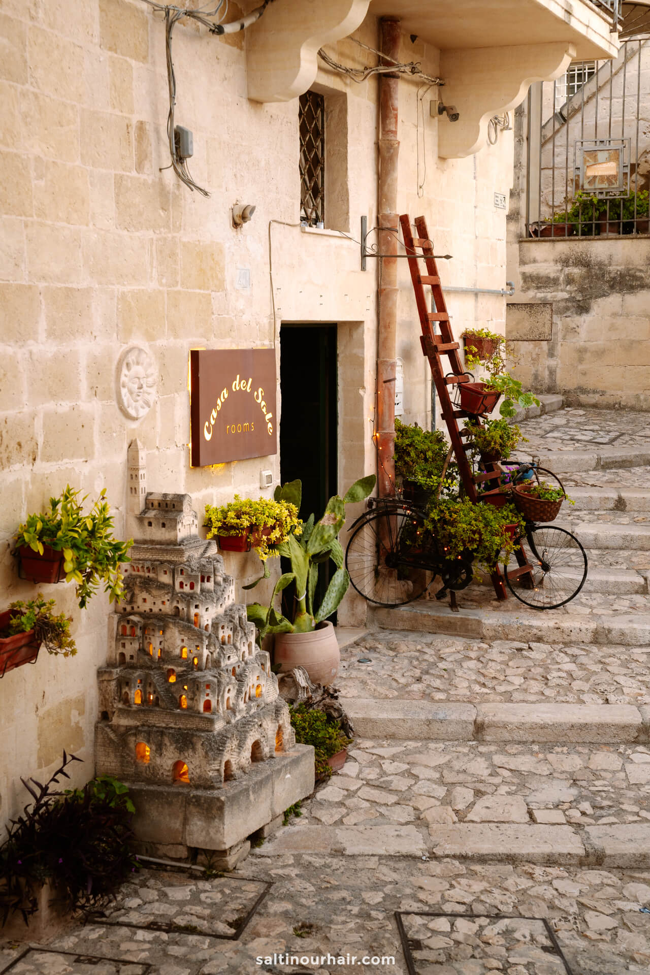 A stone-paved alley in Italy with potted plants and a decorative bicycle with a wooden ladderâ€”perfect for those wondering where to stay in Matera.