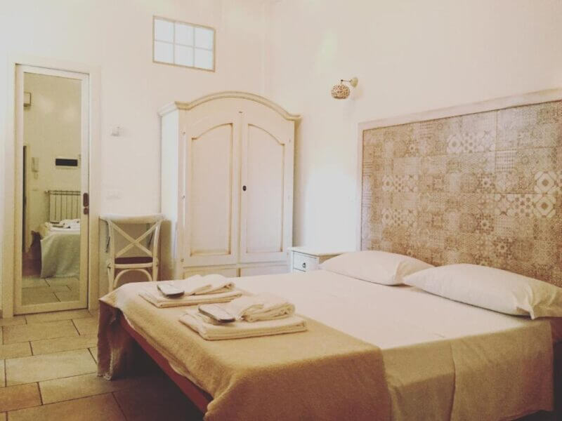 A neatly made double bed with folded towels, a patterned headboard, and a wardrobe adorn this softly lit bedroom at Donna Nina in Monopoli. A small window lets in gentle light, creating a welcoming retreat.
