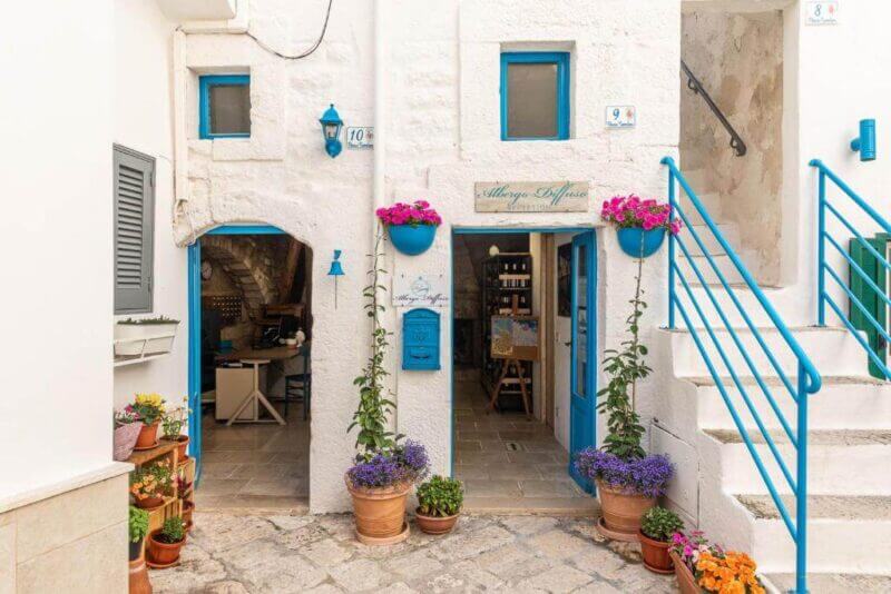 Charming white stone building with blue trim, blue door and windows, decorated with potted flowers; experience Monopoli Albergo Diffuso style with an exterior stairway on the right and a cozy seating area inside to the left.