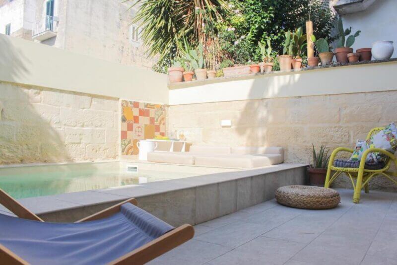 Charming Casa courtyard in Lecce featuring a rectangular pool, beige lounge seating, potted plants on the ledge, and MaHe-style wicker chair with cushion and a round woven ottoman.